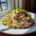 A mound of creamy salmon salad mixed with celery, red onion, and green onions served on a white plate.
