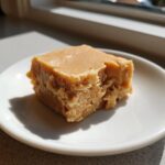 A square piece of creamy peanut butter fudge sitting on a small white plate, lit by sunlight.