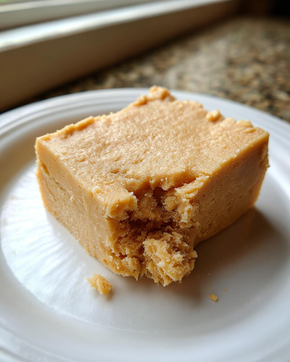 Close-up of a square of creamy, light brown peanut butter fudge on a white plate, showing a bite taken out.