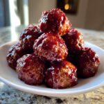 A close-up shot of a small white bowl piled high with glossy, saucy cranberry meatballs.