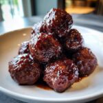 A pile of glossy, dark brown grape jelly meatballs coated in a thick, sweet sauce, served in a white bowl.