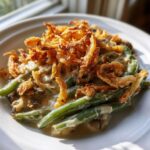 A close-up serving of amazing green bean casserole topped generously with crispy fried onions on a white plate.