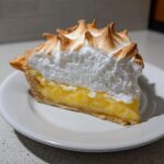 A close-up of a slice of lemon meringue pie showing bright yellow filling and tall, toasted meringue peaks.