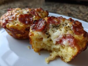 Close-up of two golden-brown pizza muffins, one with a bite taken out showing melted cheese and pepperoni topping.