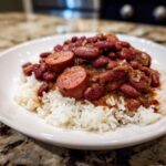 A close-up of amazing red beans and rice topped with thick slices of smoked sausage on a white plate.