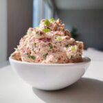 A mound of creamy, pink tuna dip mixed with visible red onion and green celery pieces in a white bowl.