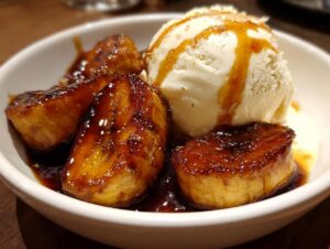 Close-up of caramelized bananas foster served in a white bowl next to a scoop of vanilla ice cream drizzled with sauce.