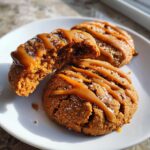 Three soft apple pie cookies on a white plate, drizzled with caramel sauce and sprinkled with sugar.