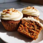 Three delicious carrot cake muffins on a white plate, one cut open showing shredded carrots inside the moist crumb.