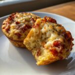 Two golden-brown pizza muffins on a white plate, one broken open to show the soft interior and melted cheese topping.