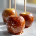 Close-up of a glossy, chewy caramel apple on a wooden stick, sitting on a white surface.