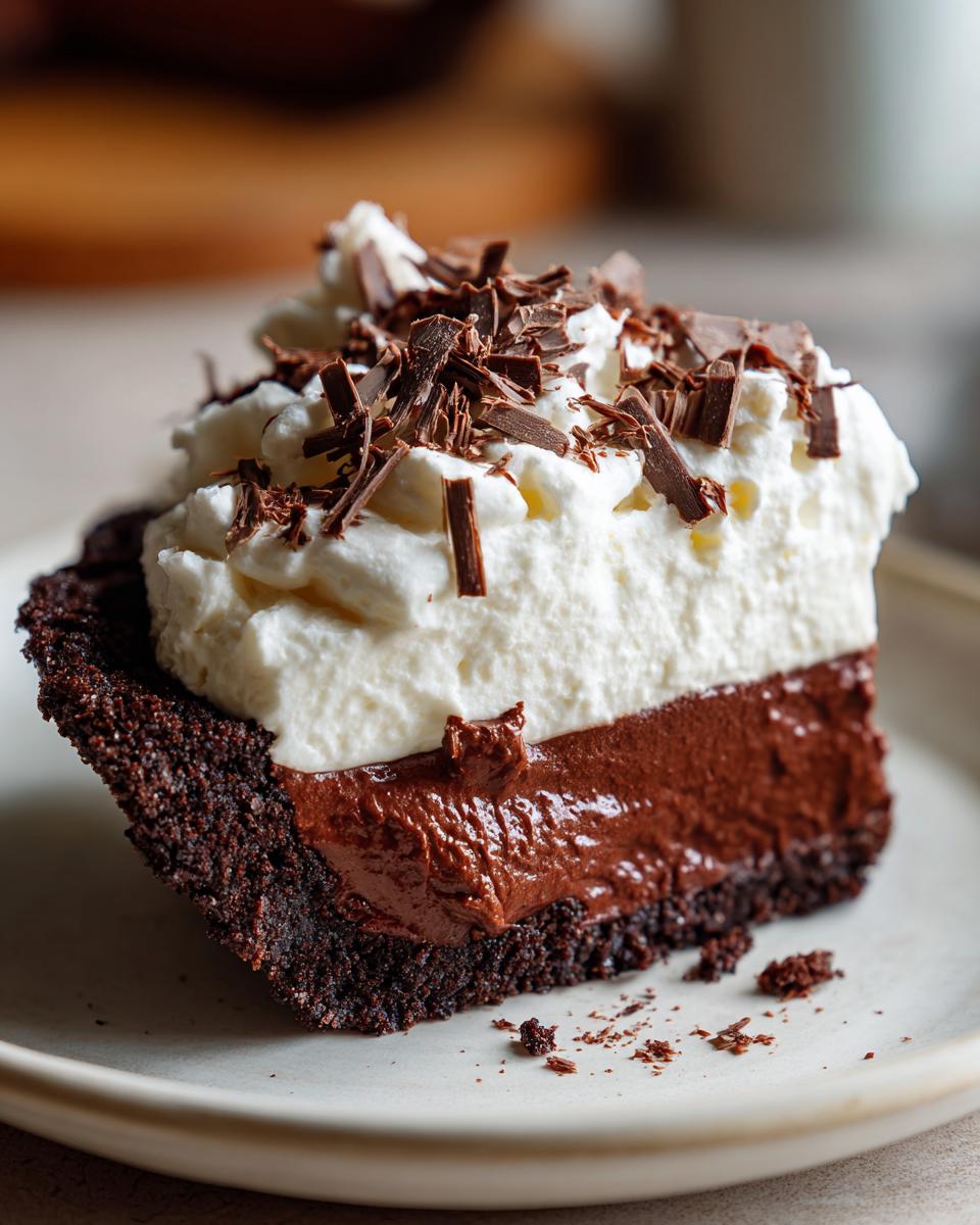 A close-up of a slice of Christmas chocolate cream pie featuring a dark crust, rich chocolate filling, and whipped cream topped with shavings.