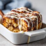 A single serving of rich Cinnamon roll casserole topped with thick white icing drizzle in a white dish.