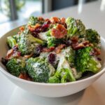 Close-up of a creamy broccoli salad topped with crispy bacon crumbles and dried cranberries in a white bowl.