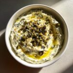 Overhead view of a bowl filled with creamy dirty martini dip, garnished with green olives, nuts, and olive oil.