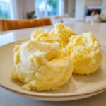 Close-up of three scoops of rich, yellow creamy mashed potatoes served on a light-colored plate.