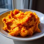 A close-up of vibrant orange, creamy mashed sweet potatoes piled high in a white bowl.