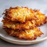 A close-up stack of three golden brown, crispy potato latkes served on a light ceramic plate.