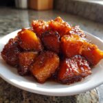 Close-up of glossy, caramelized chunks of candied yams piled on a white plate.
