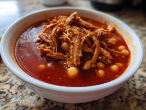 Close-up of a white bowl filled with vibrant red Pozole rojo, featuring shredded meat and hominy kernels.