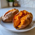 Two fluffy baked sweet potatoes on a white plate, one split open showing the bright orange interior.
