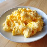 A generous serving of bright yellow, fluffy scrambled eggs piled high on a simple white plate.