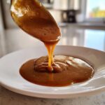 Close-up of thick, rich turkey gravy being poured from a spoon onto a white plate.