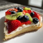 A close-up slice of fruit pizza sugar cookie topped with cream cheese frosting, strawberries, blueberries, and kiwi.