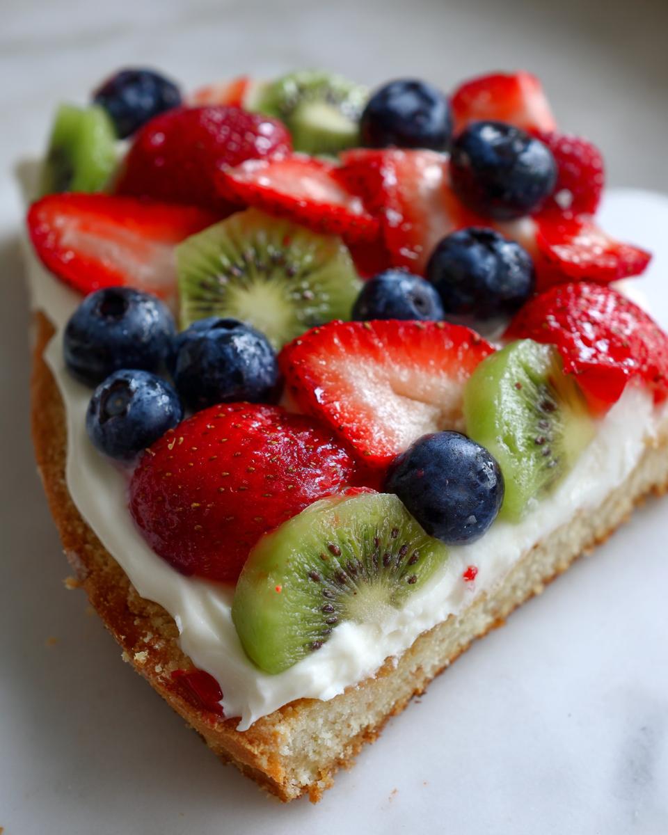 Close-up of a slice of fruit pizza sugar cookie topped with cream cheese frosting, strawberries, blueberries, and kiwi slices.