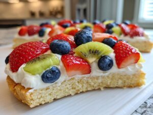 Close-up of a slice of fruit pizza sugar cookie topped with cream cheese frosting, strawberries, blueberries, and kiwi.