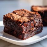 A close-up view of a rich, dark, fudgy brownies square studded with melted chocolate chips on a white plate.