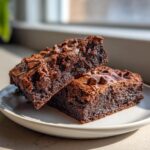 Two fudgy sweet potato brownies stacked slightly on a light plate, topped with melted chocolate chips.