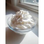 A clear glass bowl filled with stiff, glossy white whipped topping, possibly meringue or stiff royal icing recipe component.
