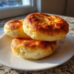 Three golden brown, fluffy cloud bread rounds stacked slightly on a white plate near a sunny window.