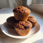 A stack of four dark, moist bran muffins with visible raisins, sitting on a white plate bathed in sunlight.