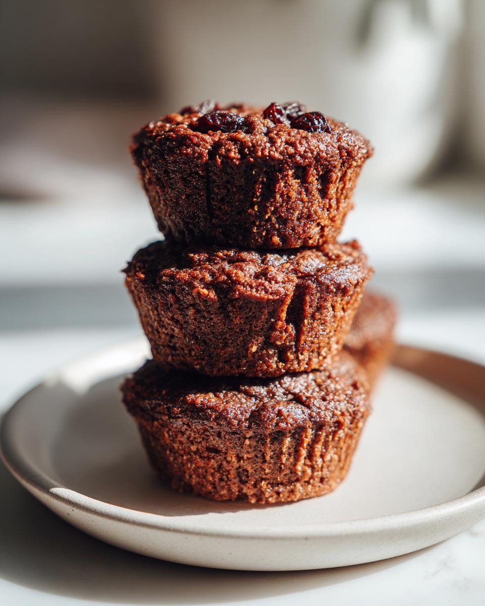 Three dark, moist bran muffins stacked on a light plate, topped with raisins, photographed in bright sunlight.