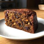 Close-up of a moist slice of dark fruit cake packed with dried fruits and nuts on a white plate.