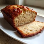 A loaf of moist greek yogurt banana bread, partially sliced, showing its tender crumb and dark, caramelized top.