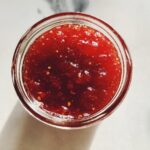 Overhead view of a glass jar filled with thick, vibrant red christmas jam, showing visible fruit pulp and seeds.