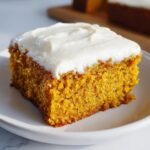 Close-up of a moist, square slice of orange pumpkin bars with thick, white cream cheese frosting.