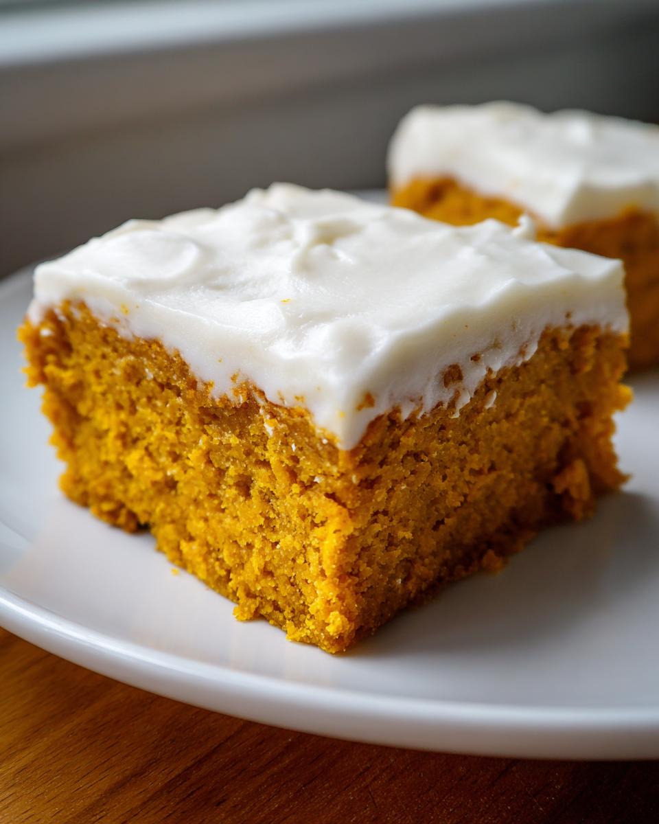A close-up of a moist, square slice of pumpkin bars with cream cheese frosting on a white plate.