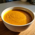 A close-up of a white bowl filled with vibrant orange roasted butternut squash soup, seasoned with pepper.