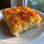A square serving of creamy hashbrown casserole topped with melted cheese and crunchy cornflakes.