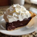 A perfect slice of rich Chocolate cream pie topped high with fluffy white whipped cream, served on a white plate.