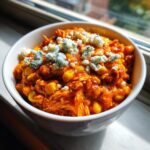 A close-up of a white bowl filled with vibrant orange slow cooker buffalo chicken chili topped with crumbled blue cheese.