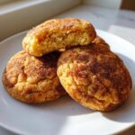 Three freshly baked Snickerdoodle cookies piled on a white plate, one broken open to show the soft interior.
