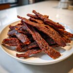 A close-up of a pile of dark, richly seasoned venison jerky strips stacked on a white plate.