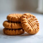 A close-up of four freshly baked peanut butter cookies stacked, featuring the classic criss-cross fork marks.