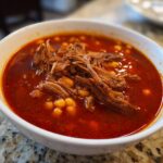 Close-up of a white bowl filled with vibrant red Pozole rojo, featuring hominy and generous amounts of shredded pork or beef.