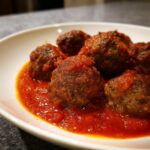 Close-up of several tender Italian meatballs submerged in rich, bright red tomato sauce in a white bowl.